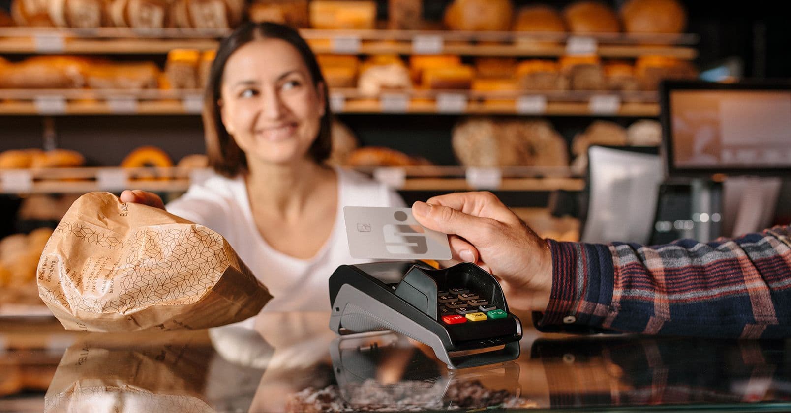 Mann bezahlt kontaktlos mit Sparkassen-Kreditkarte an einem Kartenlesegerät in einer Bäckerei, Verkäuferin reicht Tüte über den Tresen
