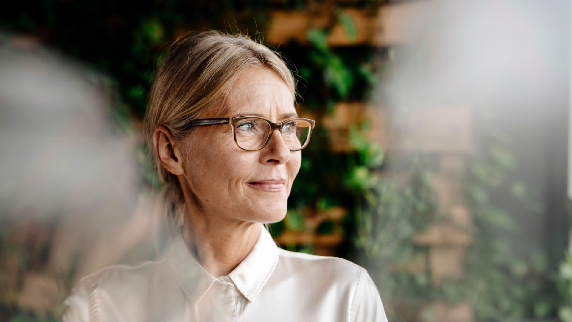 Frau mittlerem Alters schaut aus dem Fenster. Im Hintergrund ist ein begrüntes Büro erkennbar.