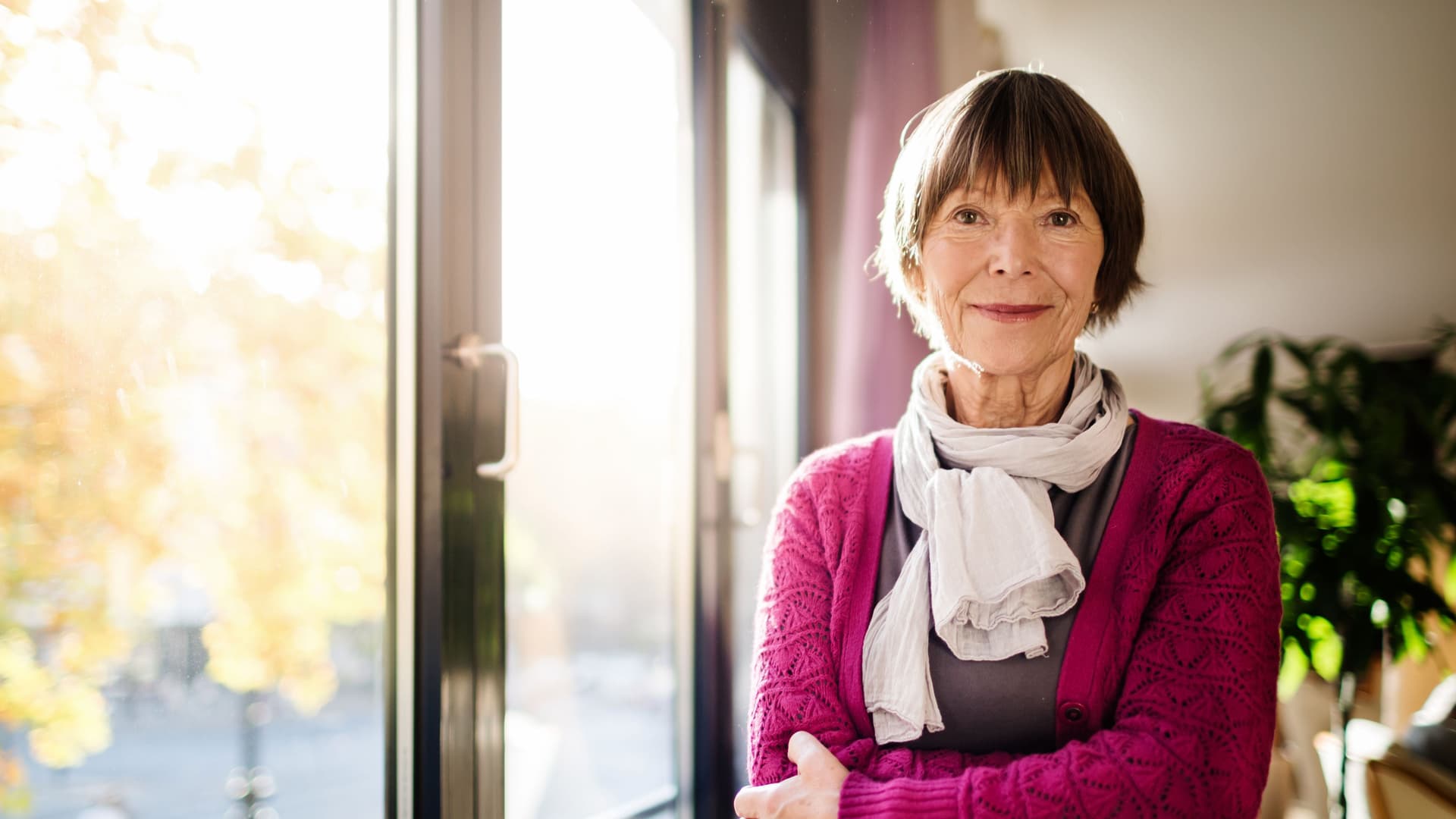 Ältere Frau, die am Fenster steht und in die Kamera lächelt. Der Blick durch das Fenster zeigt Bäume, die in Herbstfarben in der Sonne leuchten.
