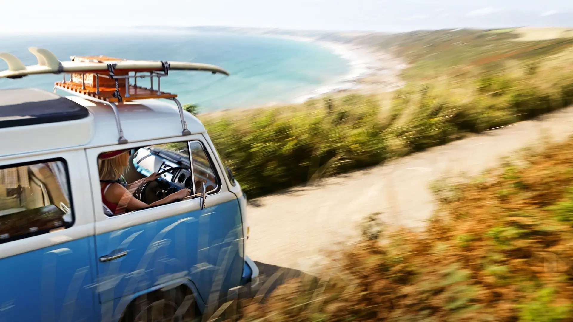 Blauer Bulli mit Surfbrett fährt auf einer sommerlichen Küstenstraße entlang