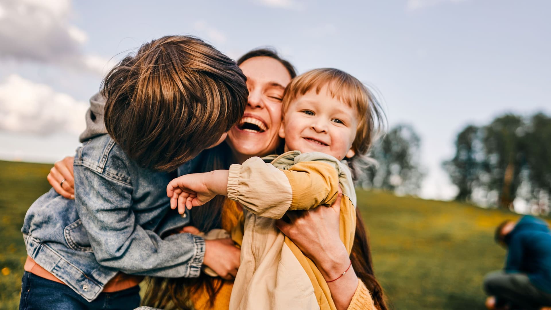 Eine junge Mutter umarmt lachend ihre zwei Kinder auf einer Wiese.