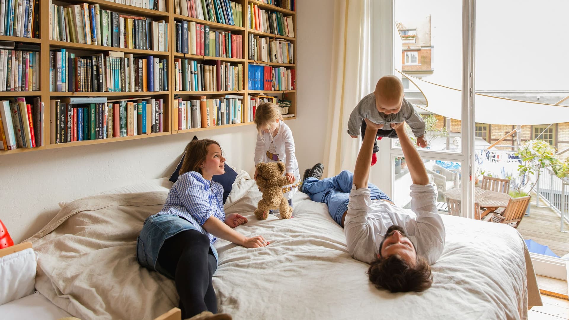 Eine vierköpfige Familie spielt zusammen auf einem Bett. Über dem Bett hängt ein großes Bücherregal.