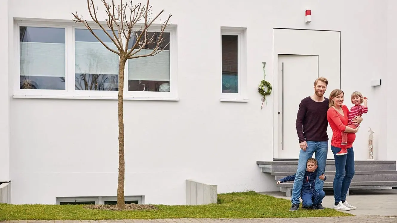 Eine schwangere Frau mit einem Baby im Arm und ein Mann mit einem Kindergartenkind stehen vor einem modernen Wohnhaus und schauen richtig Kamera.