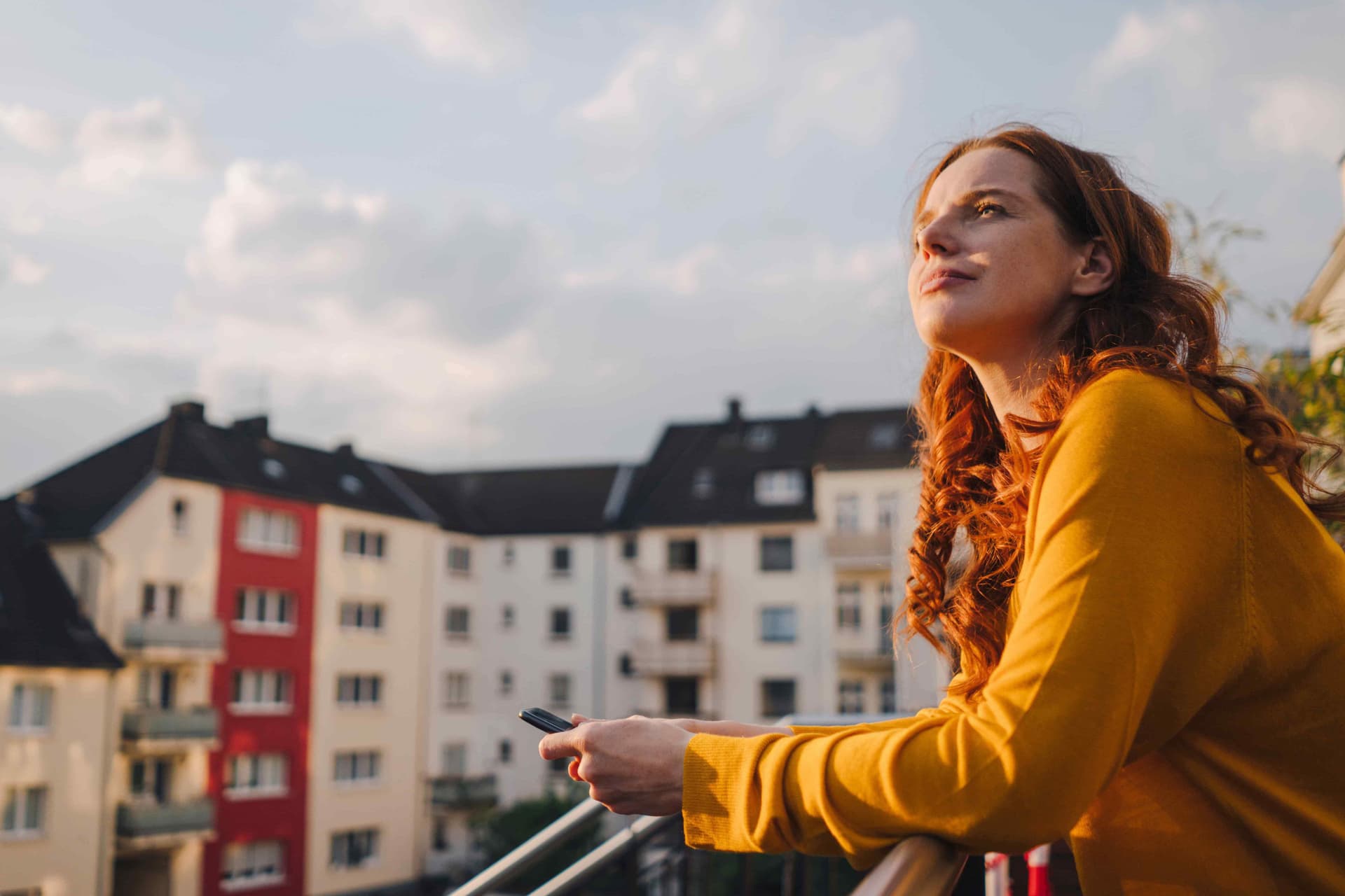 Junge Frau mit langen roten Locken steht auf einem Dachbalkon und schaut in den blauen Himmel. Im Hintergrund sind Mietswohnungen zu sehen.