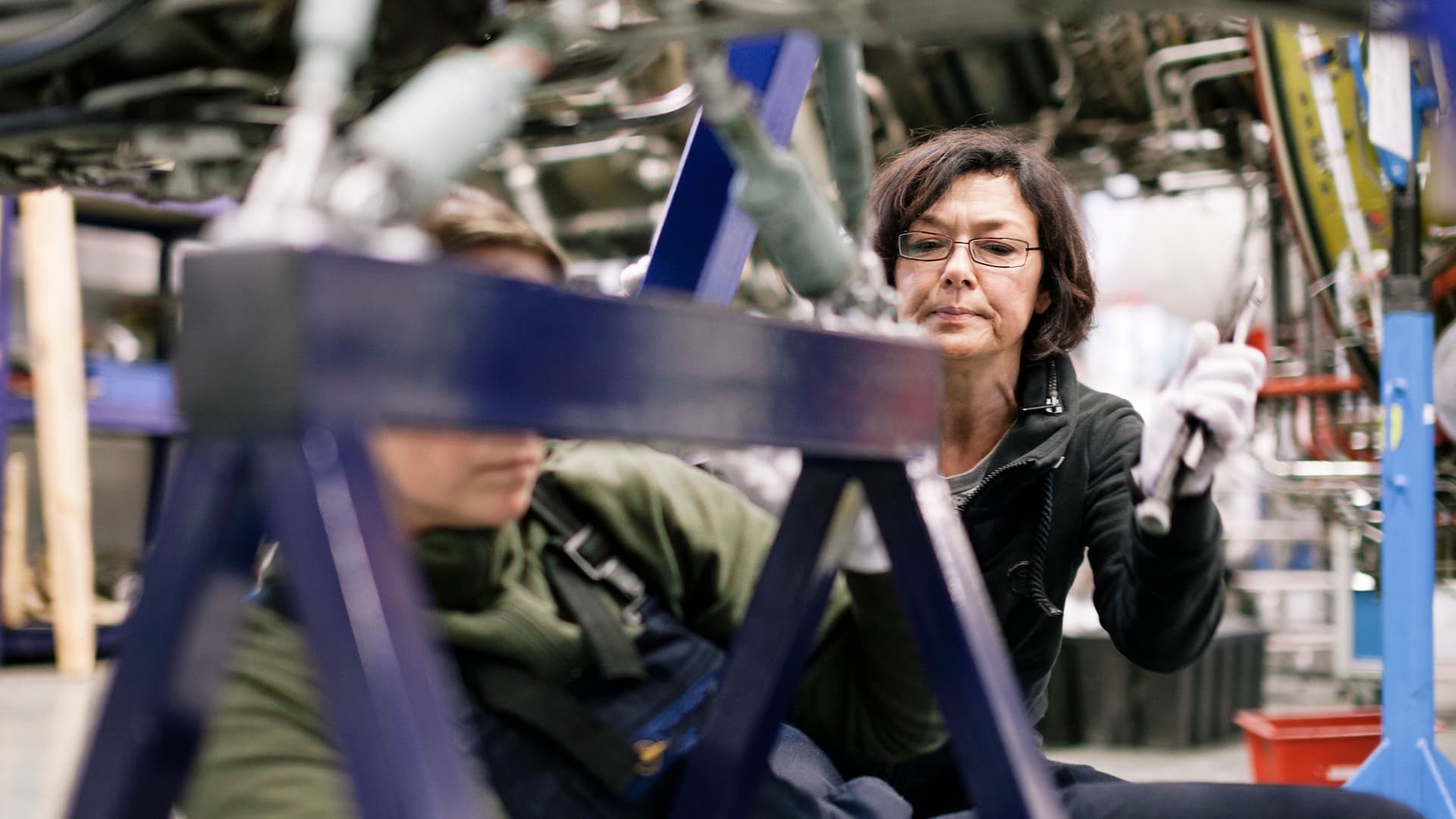 Zwei Mechanikerinnen arbeiten an einer Maschine in einem Flugzeughangar. Sie sind umgeben von Motoren und Drähten.