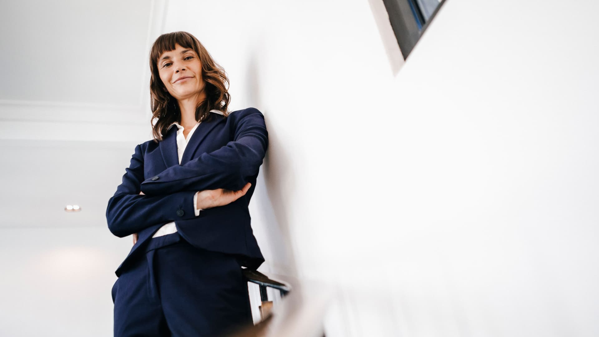 Junge Frau im Hosenanzug, die auf einer Treppe steht und die Arme verschränkt und dabei lächelt. Sie wird aus der Froschperspektive betrachtet.