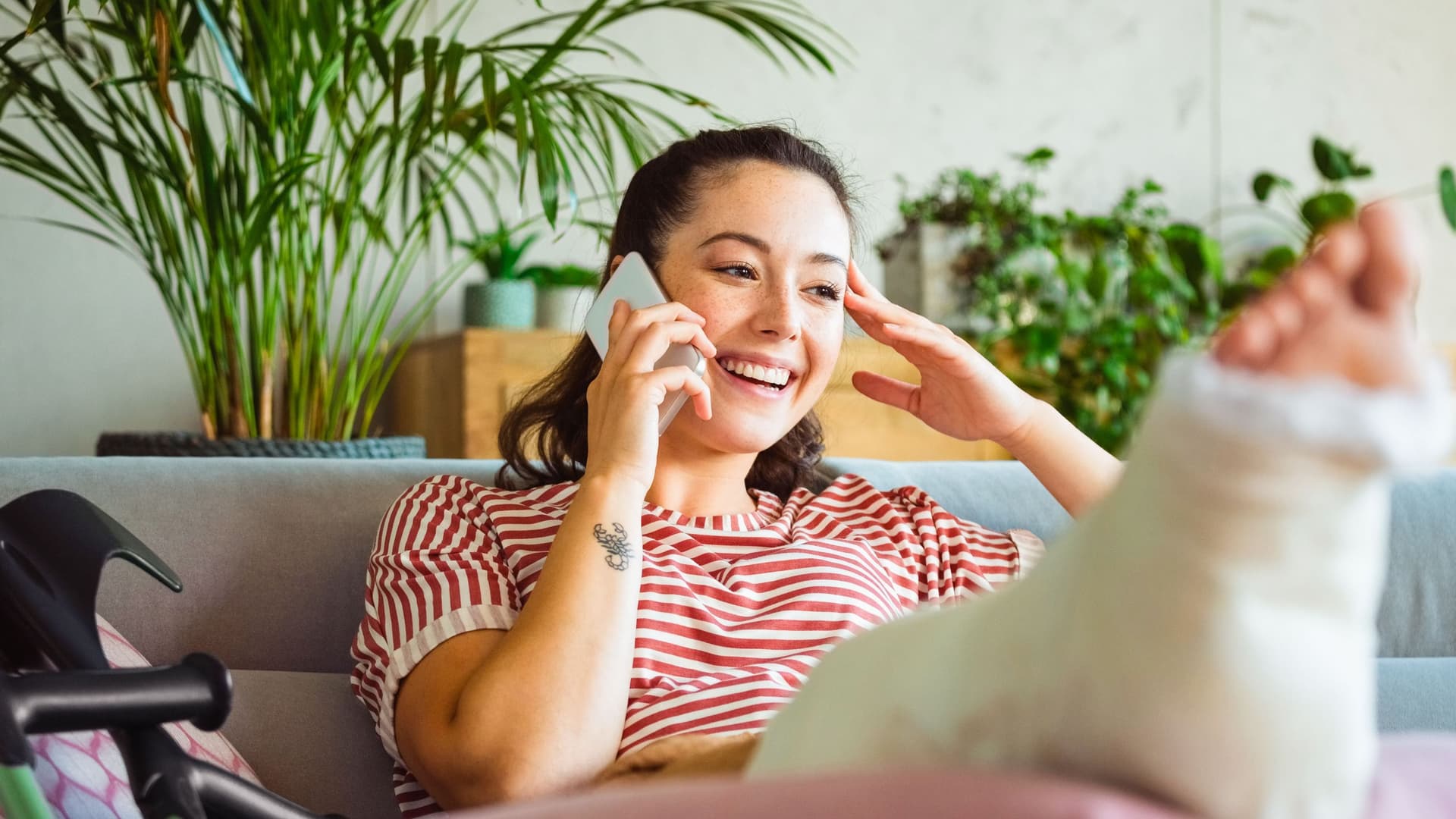 Lächelnde junge Frau mit gebrochenem Bein im Gips, die zu Hause auf dem Sofa liegt und mit dem Smartphone telefoniert.