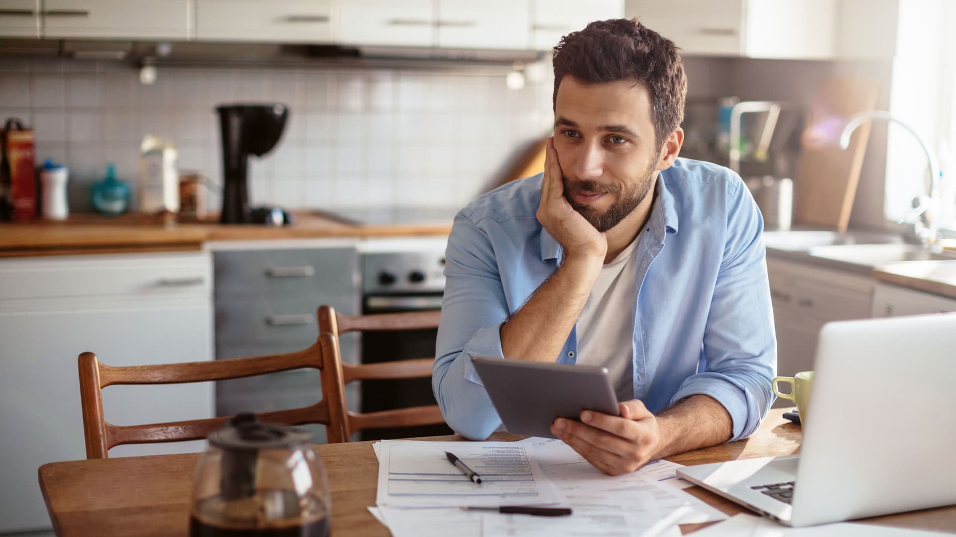 Junger Mann sitz in einer Küche mit einem Laptop am Tisch. Er schaut nachdenklich und hat ein Tablett in der Hand und Papiere vor sich.