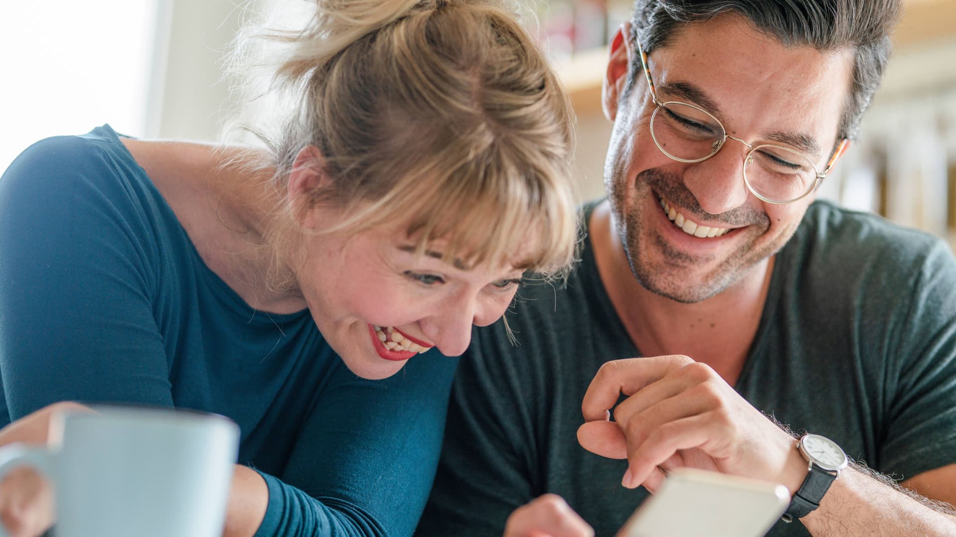 Ein Paar mittleren Alters sitzt zusammen und schaut auf ein Smartphone. Sie lachen ausgelassen.