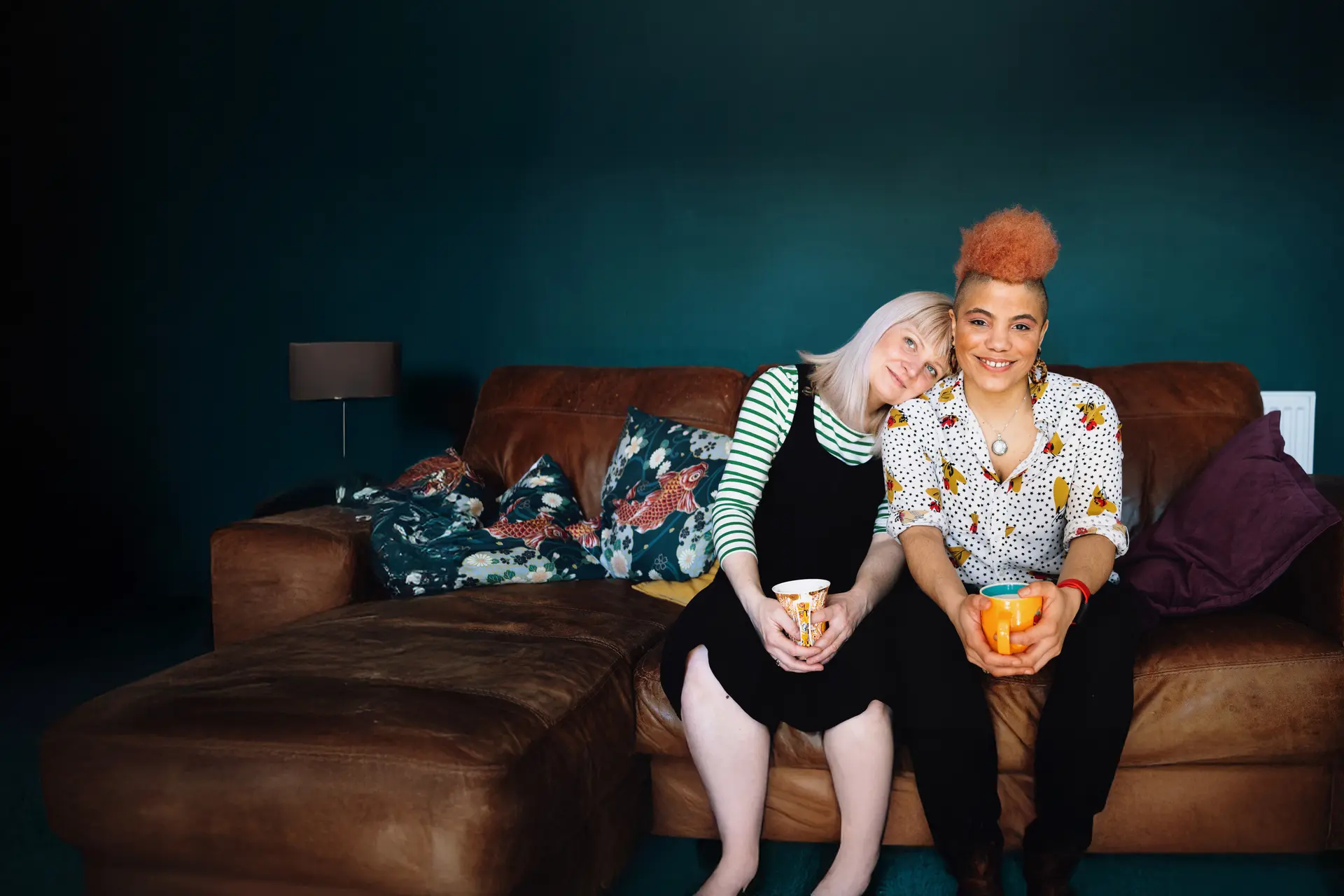 Porträt von zwei Frauen. die nebeneinander mit jeweils einer Tasse in den Händen auf einem braunen Ledersofa sitzen. Die eine Frau hat den Kopf auf die Schulter der anderen gelegt.