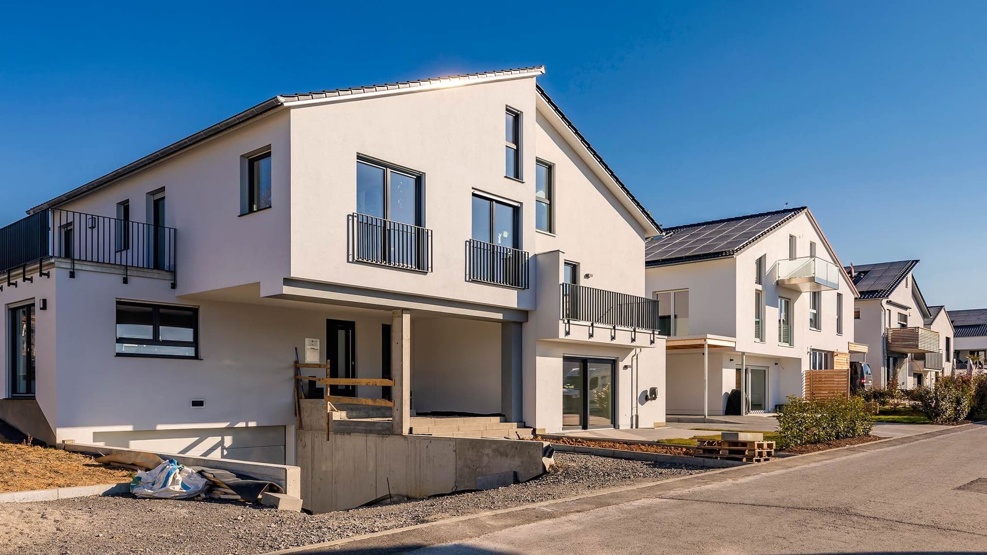 Eine Außenaufnahme eines Straßenzuges mit neugebauten Einfamilienhäusern. Ein weißes Haus ist zentral im Bild. Ähnliche Häuser reihen sich anhand einer Fluchtlinie hintereinander.