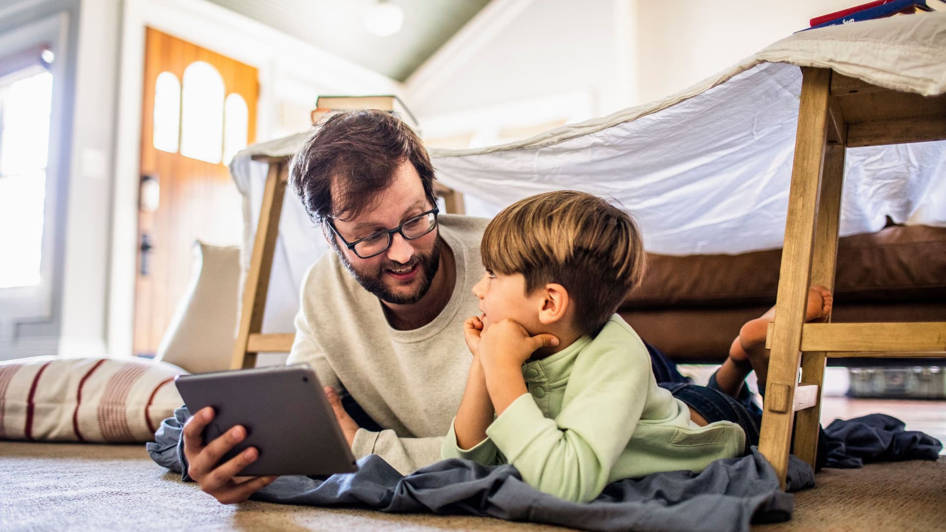 Mann und Kind liegen zusammen mit einem Tablet auf dem Boden. Über ihnen ist ein Bettlaken über zwei Stühlen gespannt.