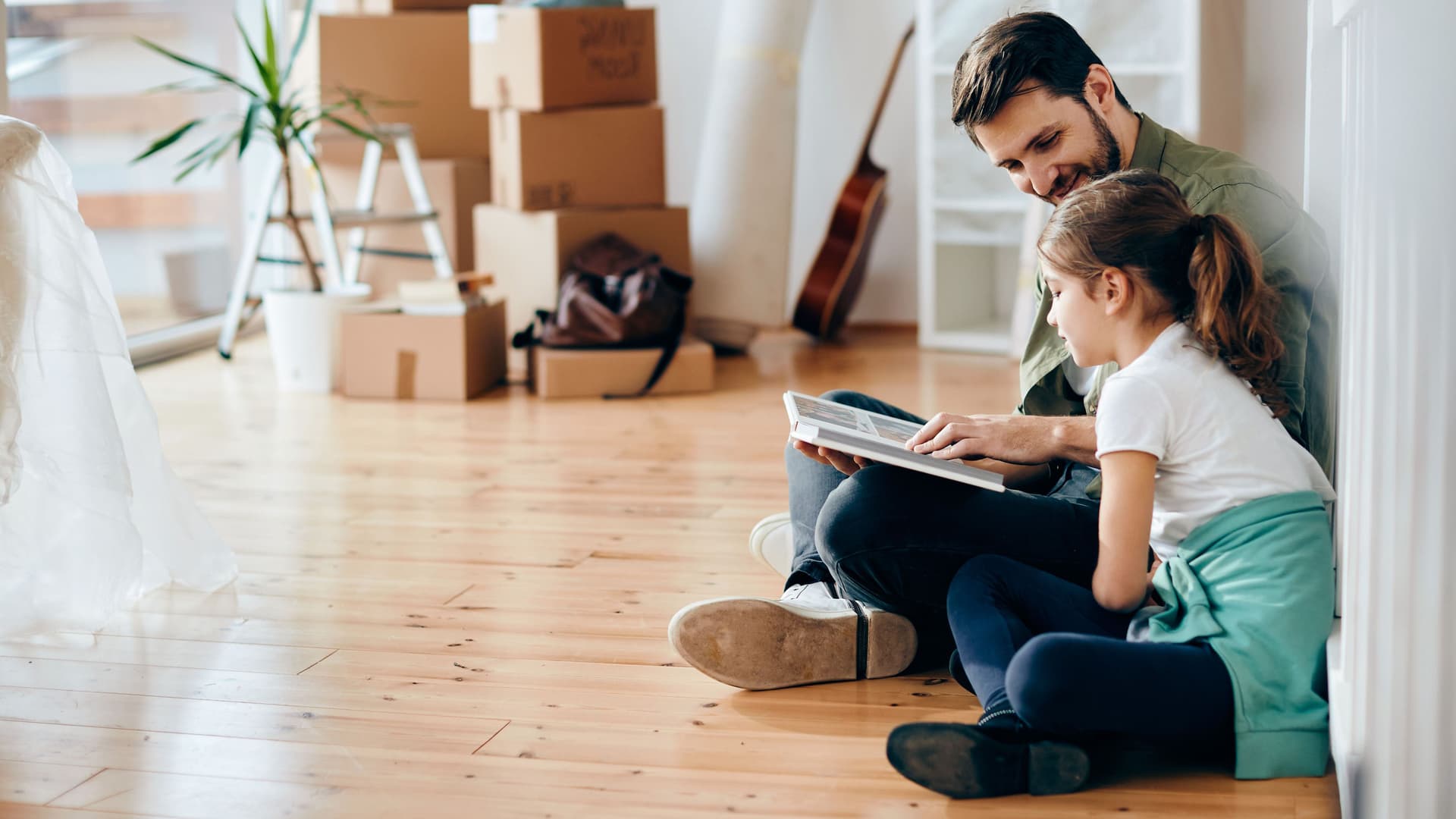 Lächelnder Vater und kleine Tochter sitzen auf dem Holzboden und gucken ein Fotoalbum an. Der Raum ist fast leer und im Hintergrund sind Umzugskisten.