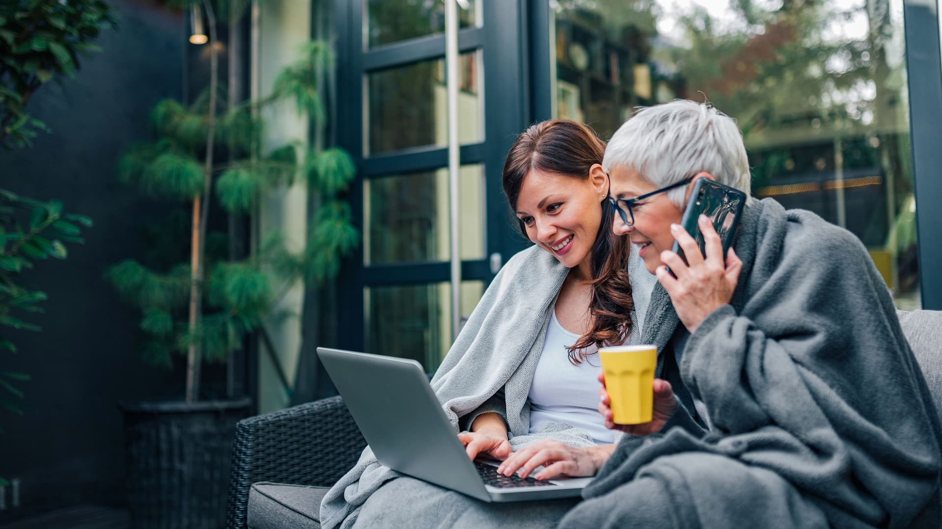 Zwei Frauen sitzen auf einer Terasse. Die eine hat ein Handy am Ohr, die andere einen Laptop auf dem Schoß.