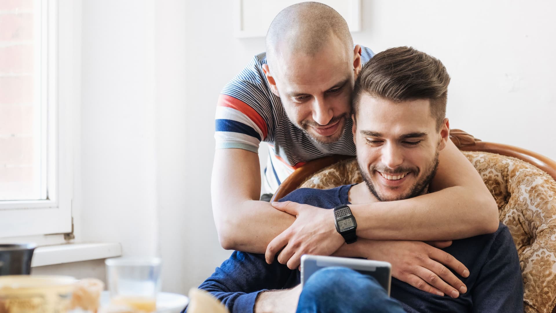Zwei Maenner kuscheln beim Frühstueck und schauen gemeinsam auf ein Tablet