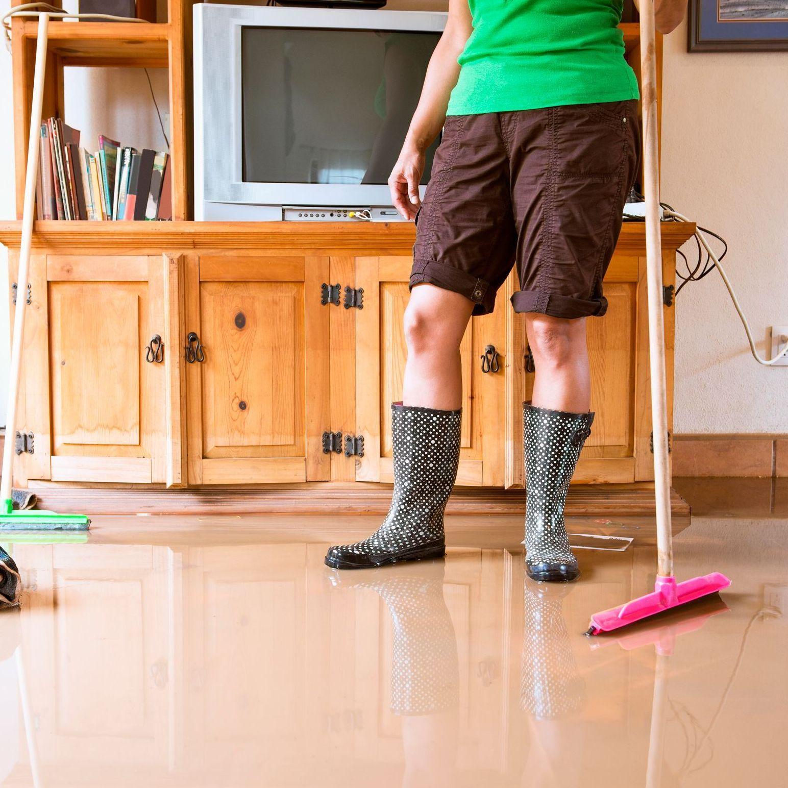 Eine Person in Gummistiefeln steht in einem überschwemmten Wohnzimmer und hält einen Besen. 