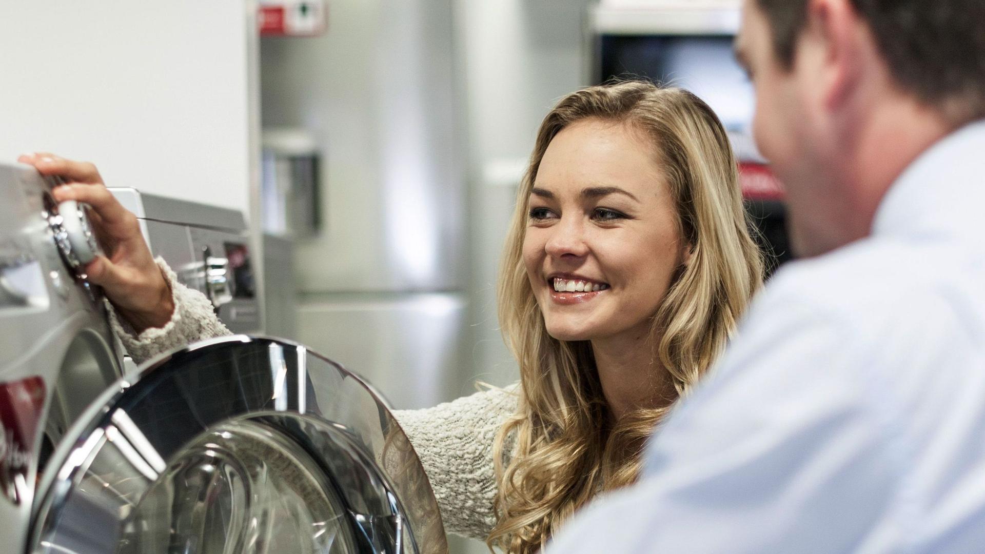 Eine junge blonde Frau ist in einem Beratungsgespräch mit einem Verkäufer. Sie hocken vor einer Waschmaschine.
