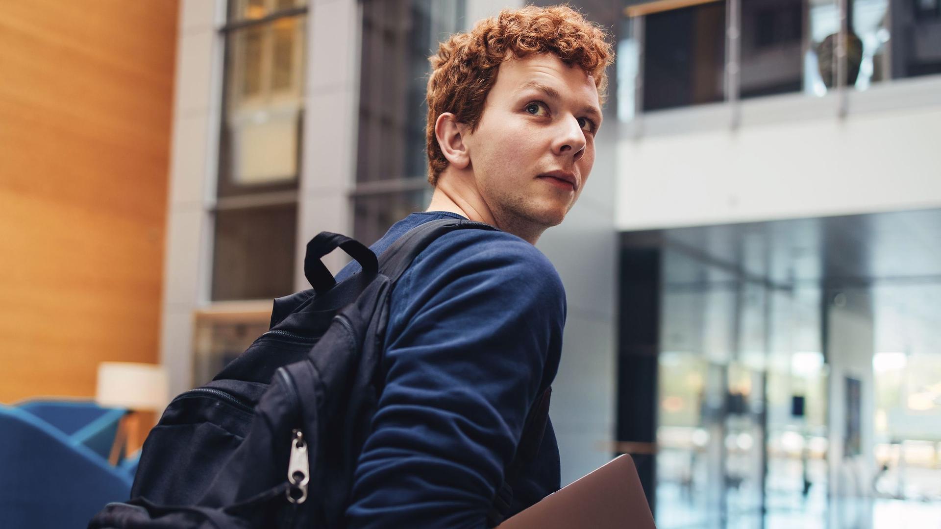 Student mit Laptop unterm Arm blickt über seine Schulter Richtung Kamera.