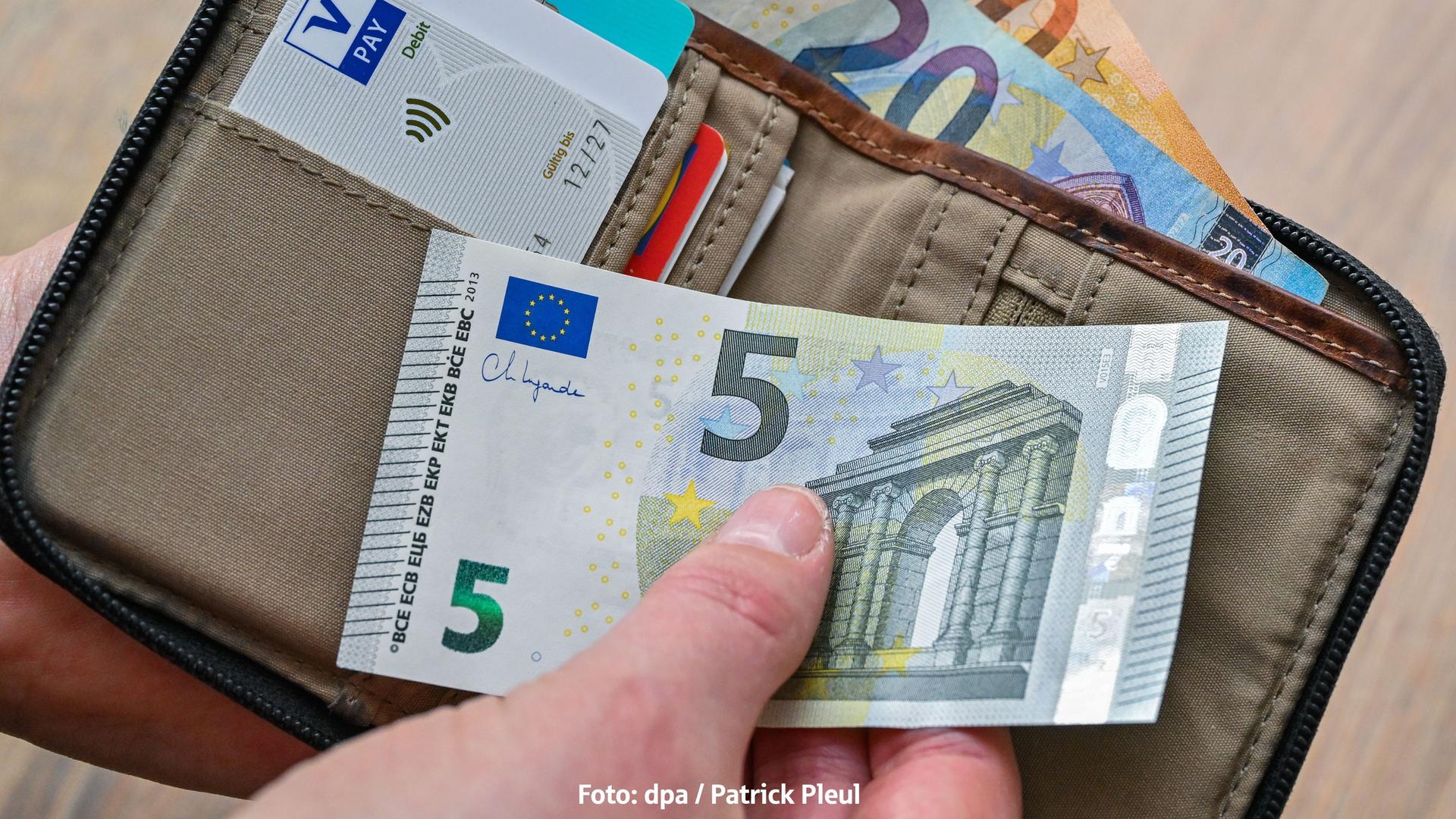 Eine Hand hält einen 5-Euro-Schein. Dahinter liegt ein Geldbeutel mit einem 20-Euro Schein und einem 50-Euro Schein.