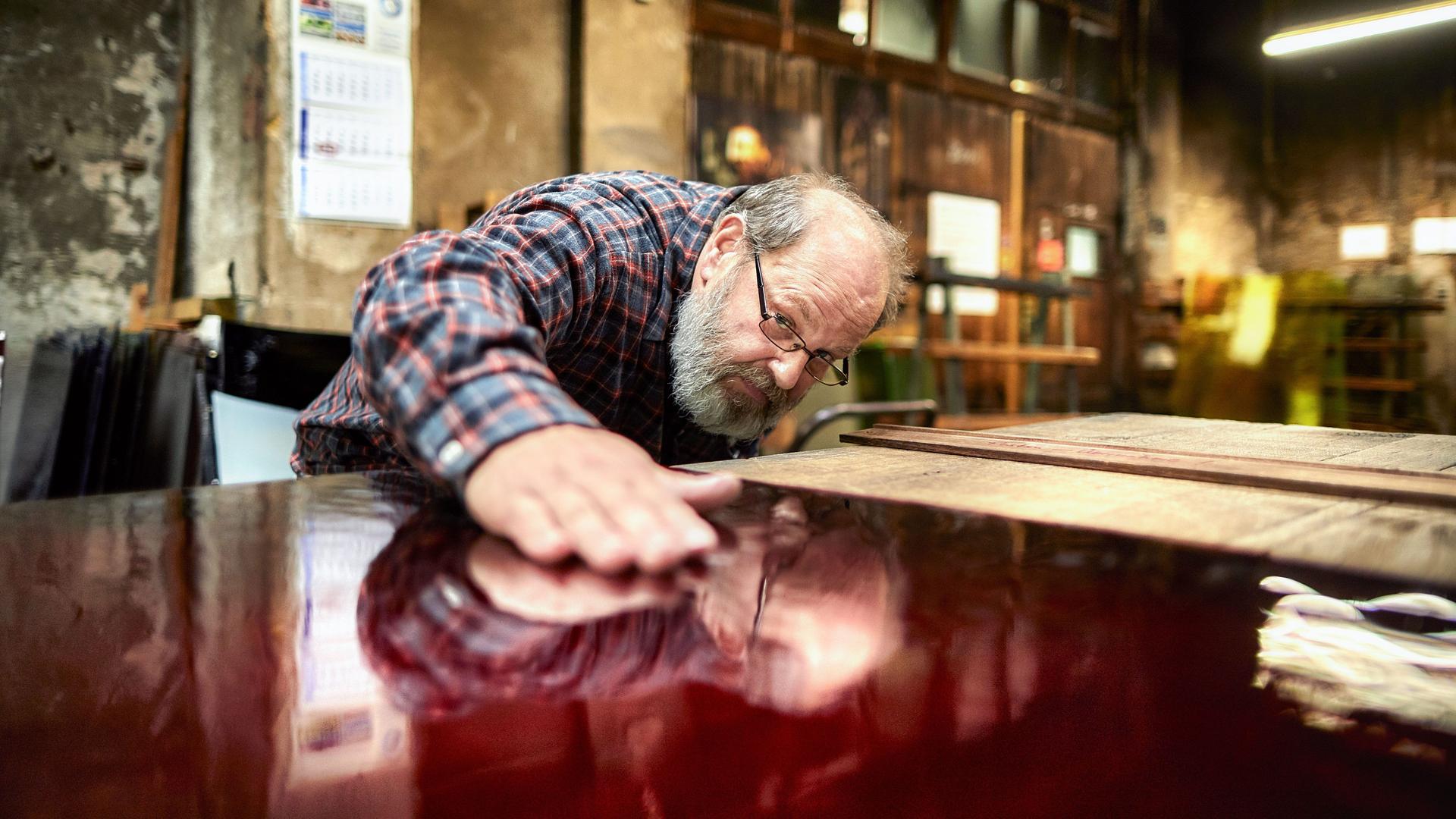 Ein älterer Mann mit Brille und Vollbart beugt sich in einer Werkstatt über eine glatte, rot lackierte Oberfläche und hält die flache Hand darauf.