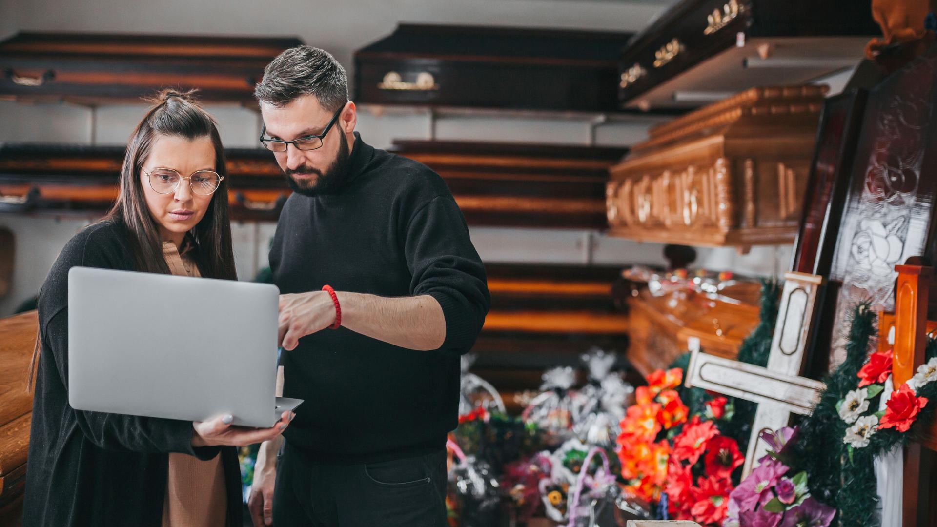 Eine Frau und ein Mann stehen mit einem Laptop vor einem Regal mit Särgen und Bestattungsartikeln.