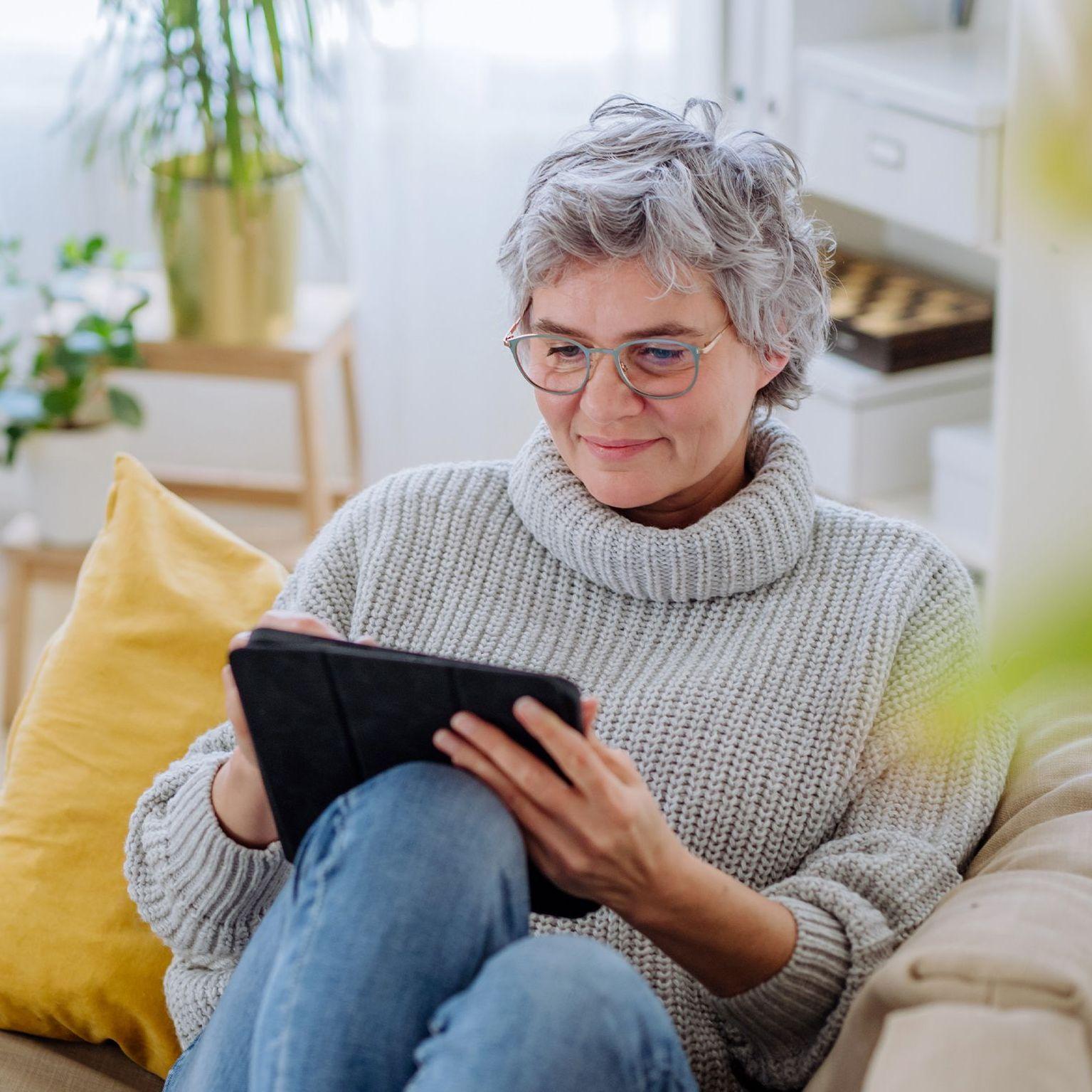 Eine Frau mit kurzen grauen Haaren sitzt zuhause bequem auf dem Sofa und schaut auf ein Tablet.
