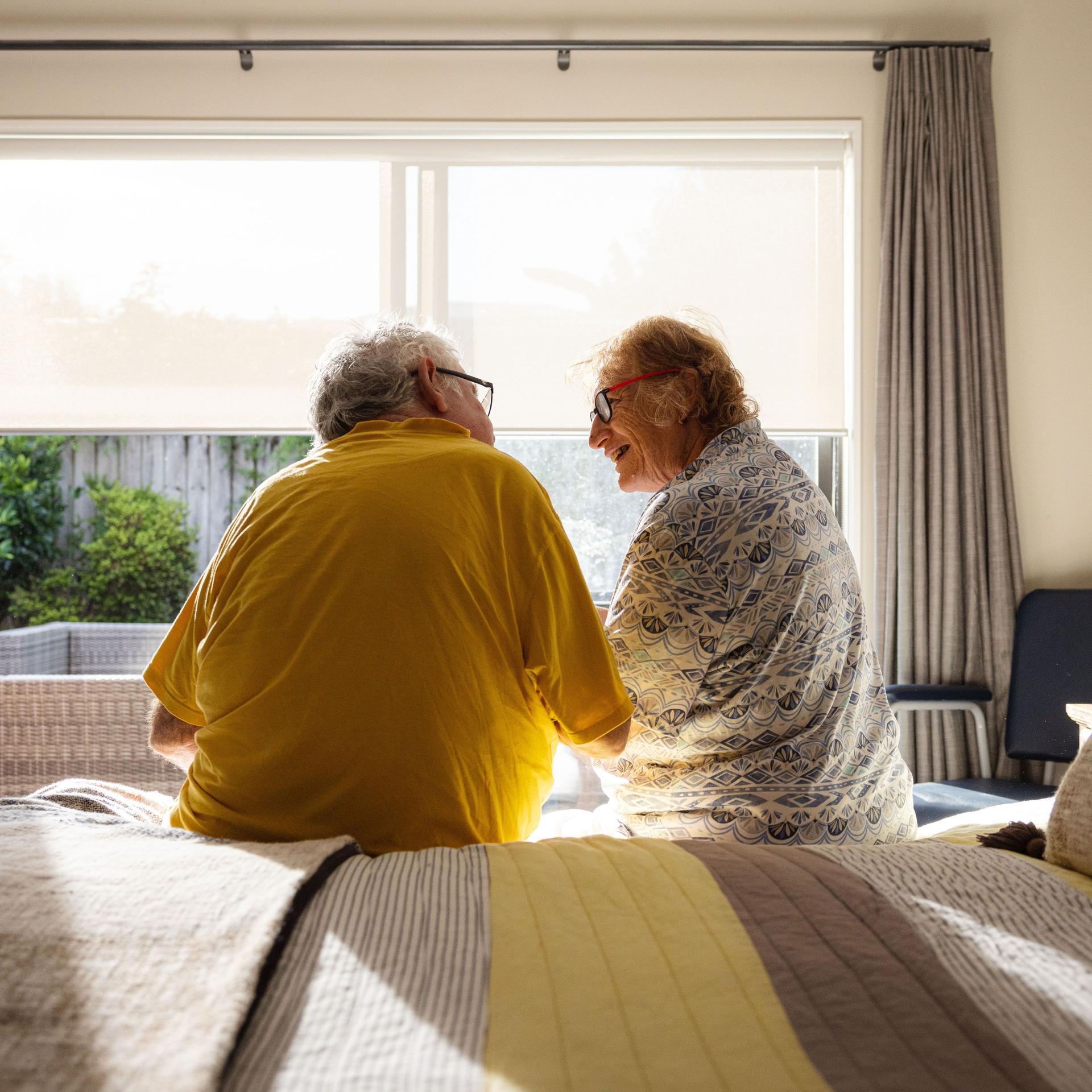 Ein älteres Paar sitzt auf dem Bett. Es scheint die Sonne. Sie lächeln sich an.
