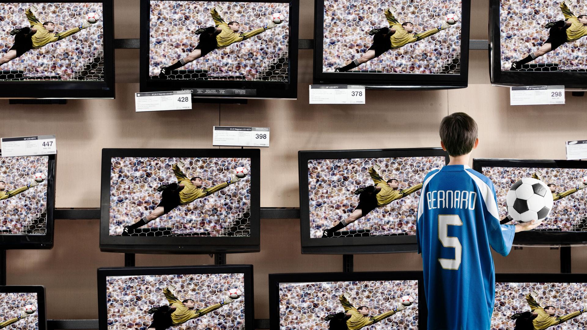 Ein Jungs mit einem Fußballtrikot an und einem Fußball in der Hand steht vor Fernsehern in einem Laden.