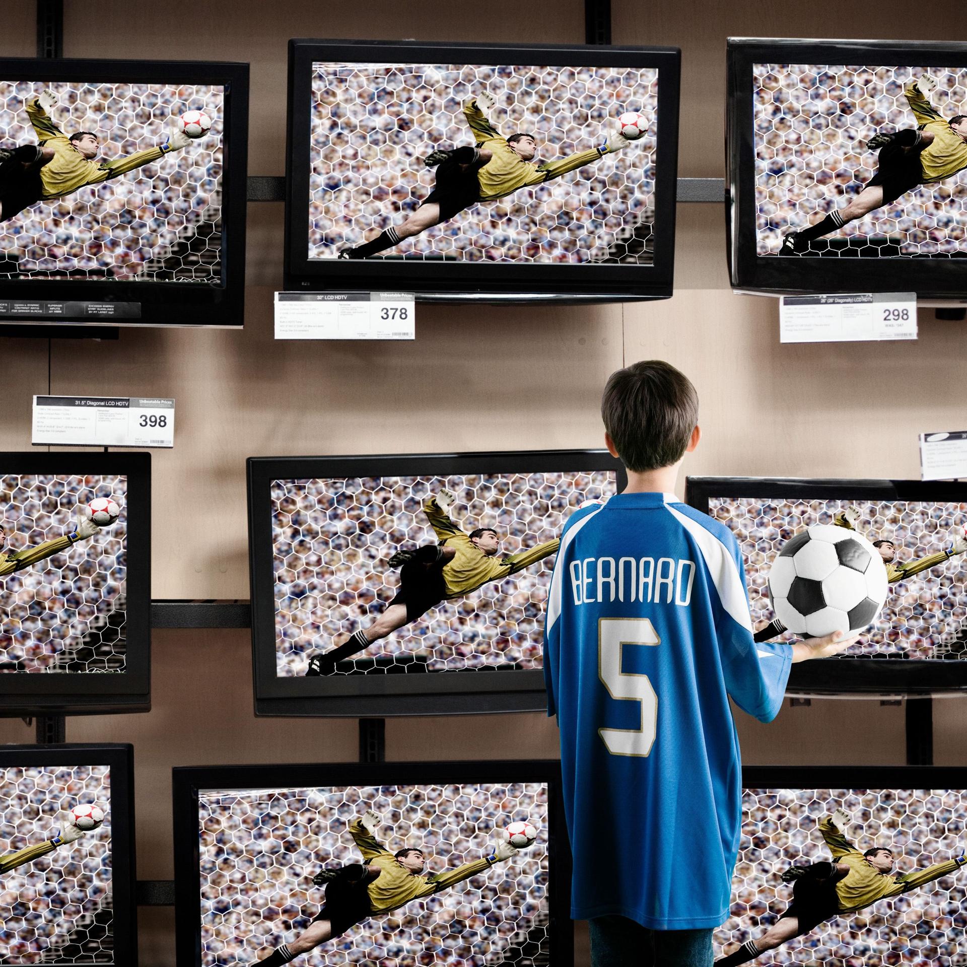 Ein Jungs mit einem Fußballtrikot an und einem Fußball in der Hand steht vor Fernsehern in einem Laden.