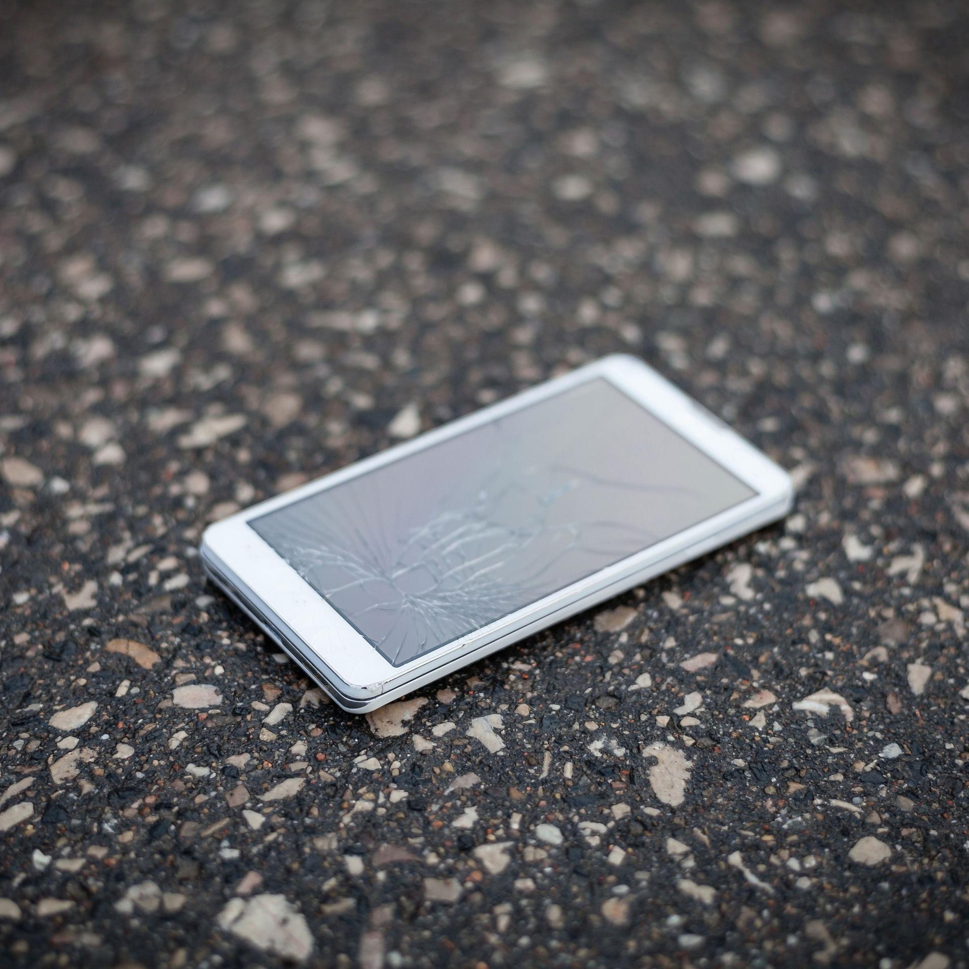 Ein weißes Smartphone liegt auf Betonboden. Die Scheibe ist gesplittert.