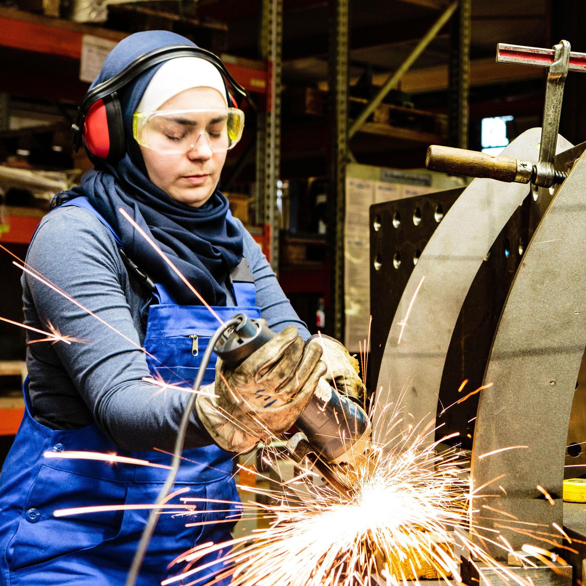 Eine junge Frau mit Kopftuch steht in einer Metall-Werkstatt. Sie arbeitet an einem Werkstück.