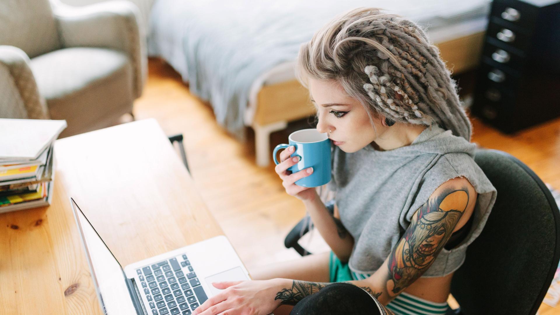 Eine junge tätowierte Frau mit grauen Dreadlocks sitzt konzentriert mit einer Tasse Kaffee vor ihrem Laptop.