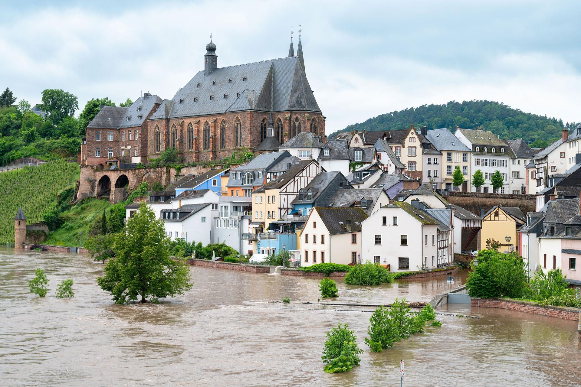 Ein überflutete Stadt am Fluss. Auf einem Hügel steht eine Kirche. Davor befinden sich überflutete Wohnhäuser.