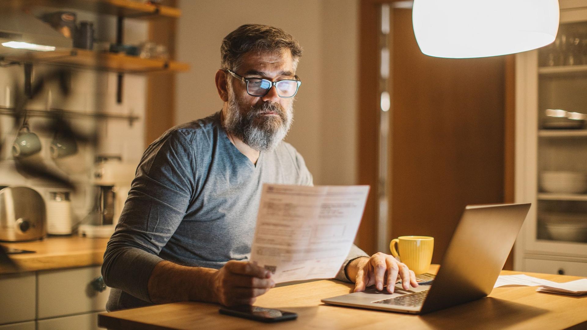 Ein Mann mittleren Alters sitzt in seiner Küche am Tisch mit seinem Laptop und liest konzentriert ein Dokument. 