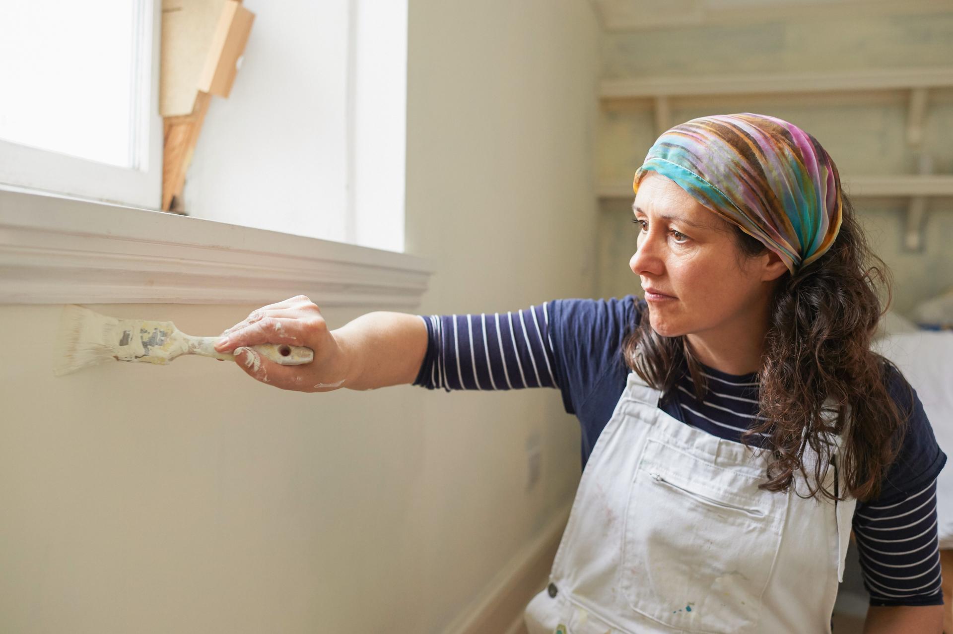 Frau in Latzhose streicht sitzend mit einem Pinsel eine weiße Wand unterhalb einer Fensterbank.