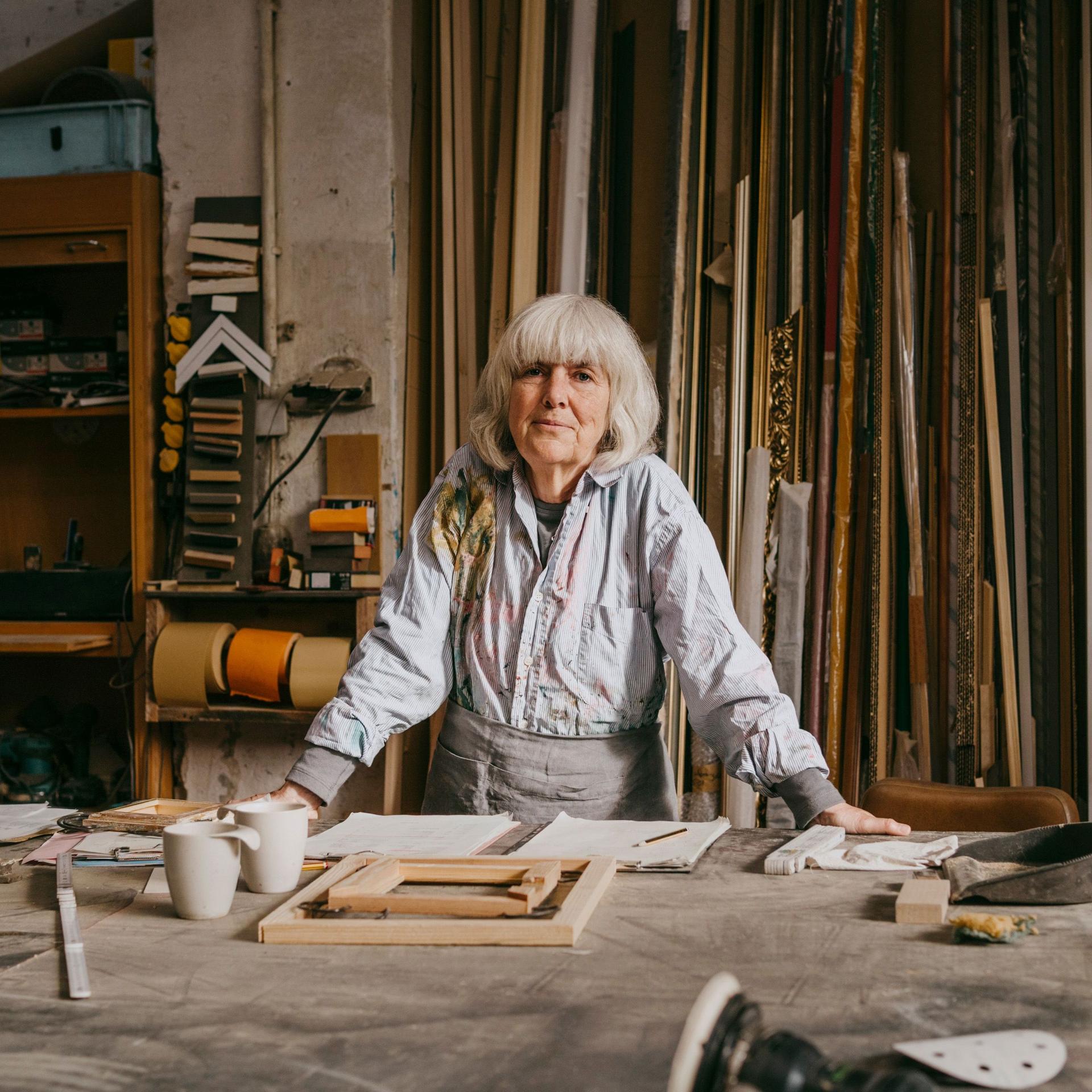 Eine Frau mit grauen Haaren steht an einem Tisch in einer Schreinerei. Im Hintergrund steht Holz.