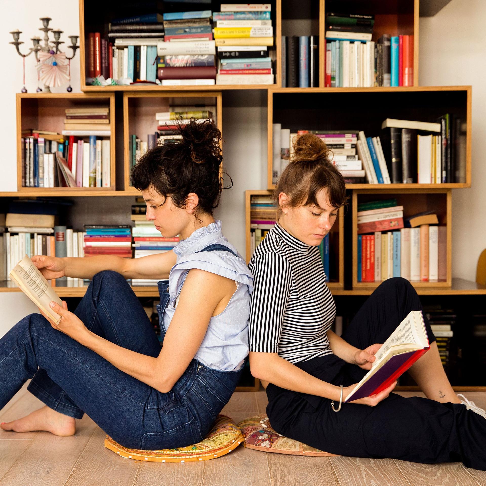 Zwei Freundinnen sitzen lesend Rücken an Rücken auf dem Boden vor einem Bücherregal