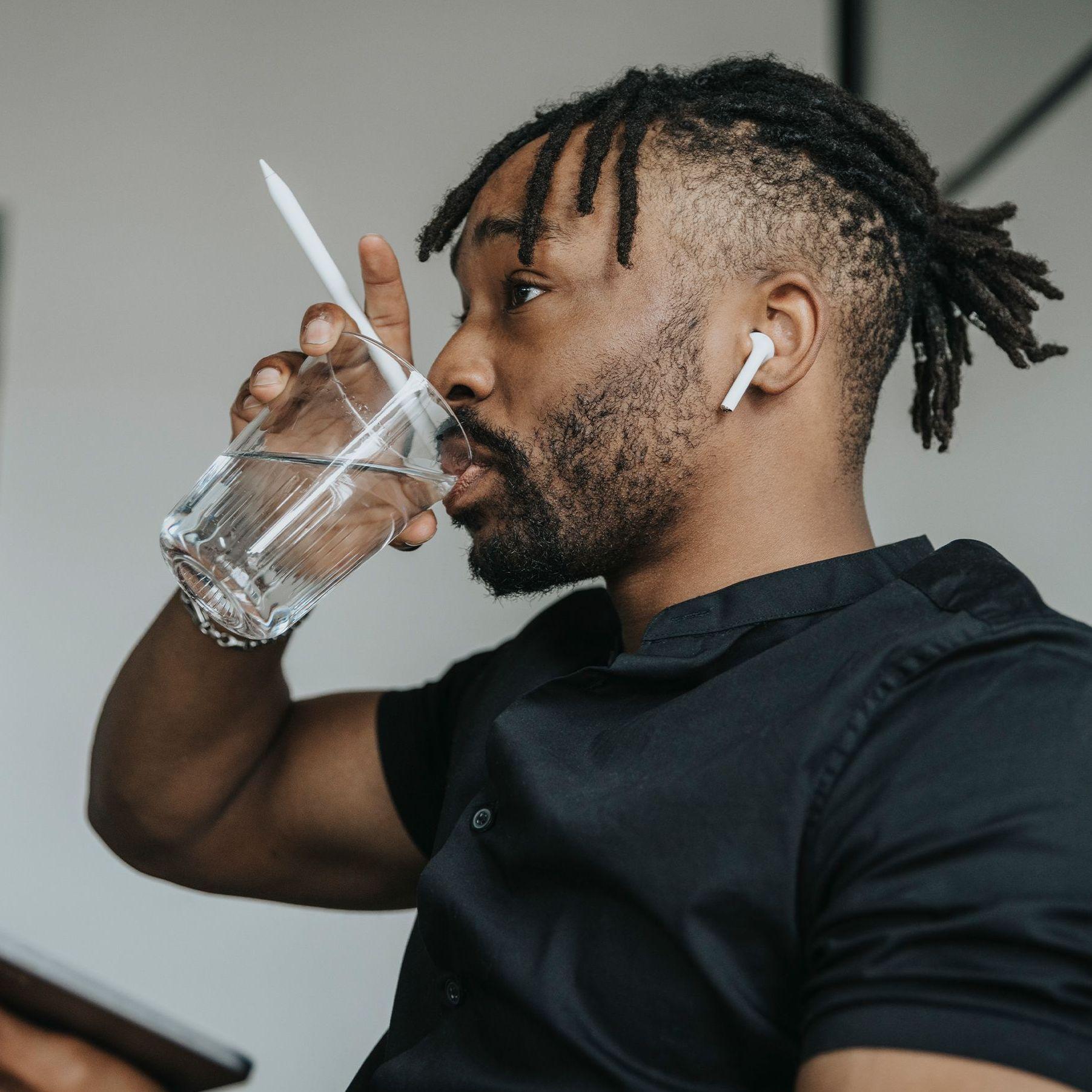 Ein junger Mann in einem kurzärmeligen T-Shirt trinkt im Büro aus einem Glas Wasser während er ein Tablet mit Stift in der Hand hält.