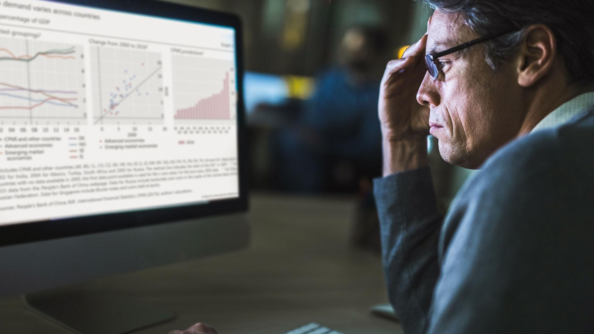 Mann mittleren Alters sitzt vor Computerbildschirm mit Statistik-Grafiken und stütz seinen Kopf mit der Hand.