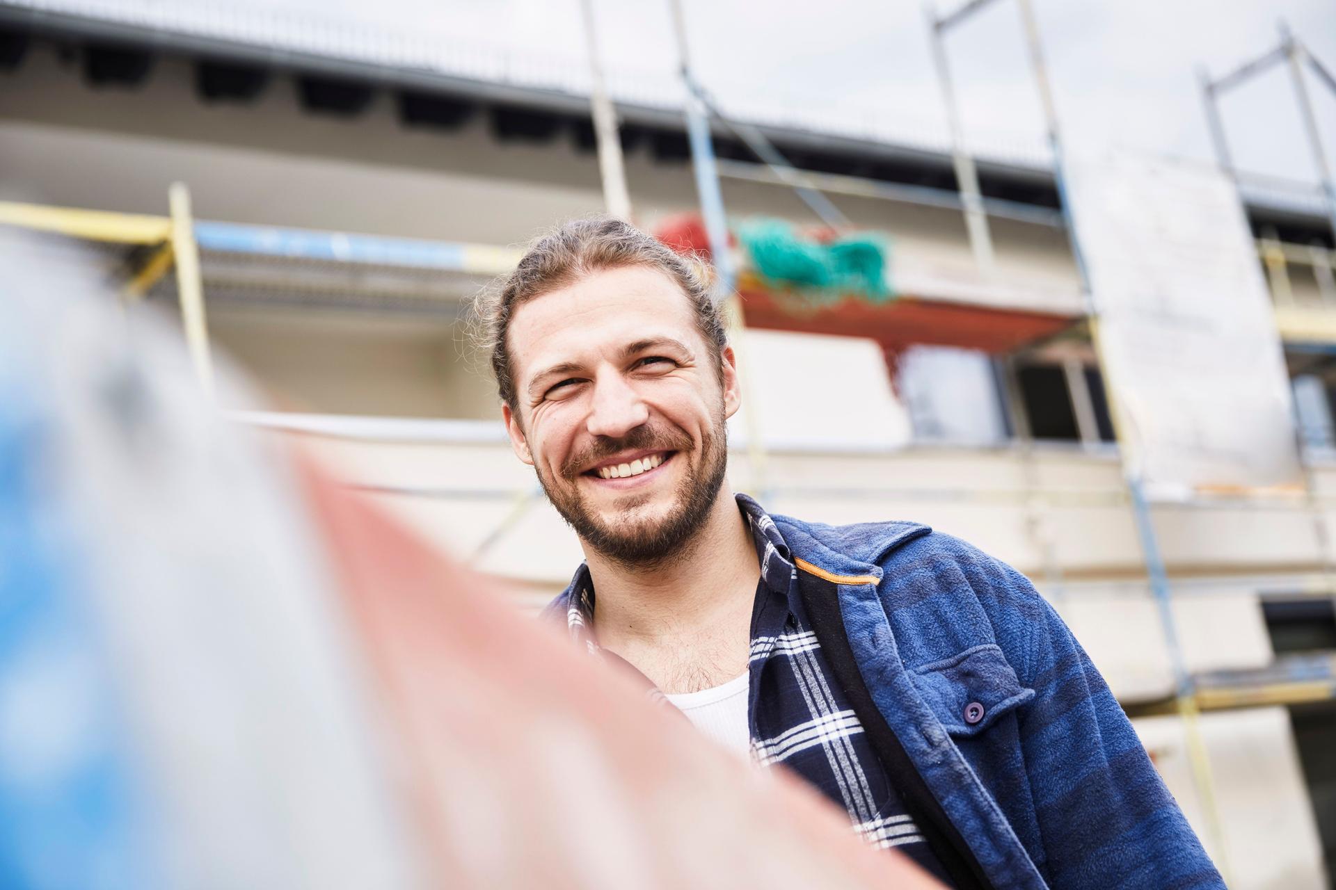 Ein junger Mann steht auf einer Baustelle und lächelt in die Kamera.