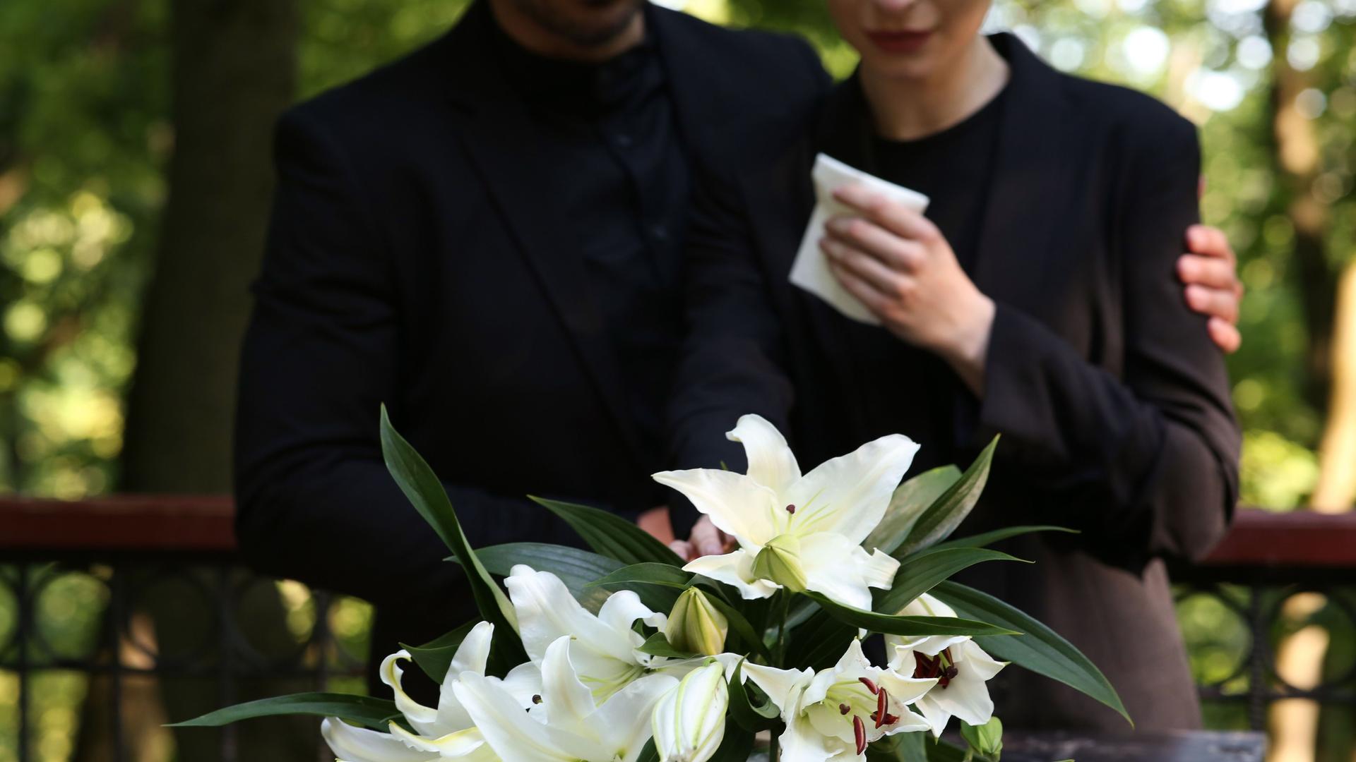 Paar in der Nähe von Granitgrabstein mit weißen Lilien auf dem Friedhof im Freien.