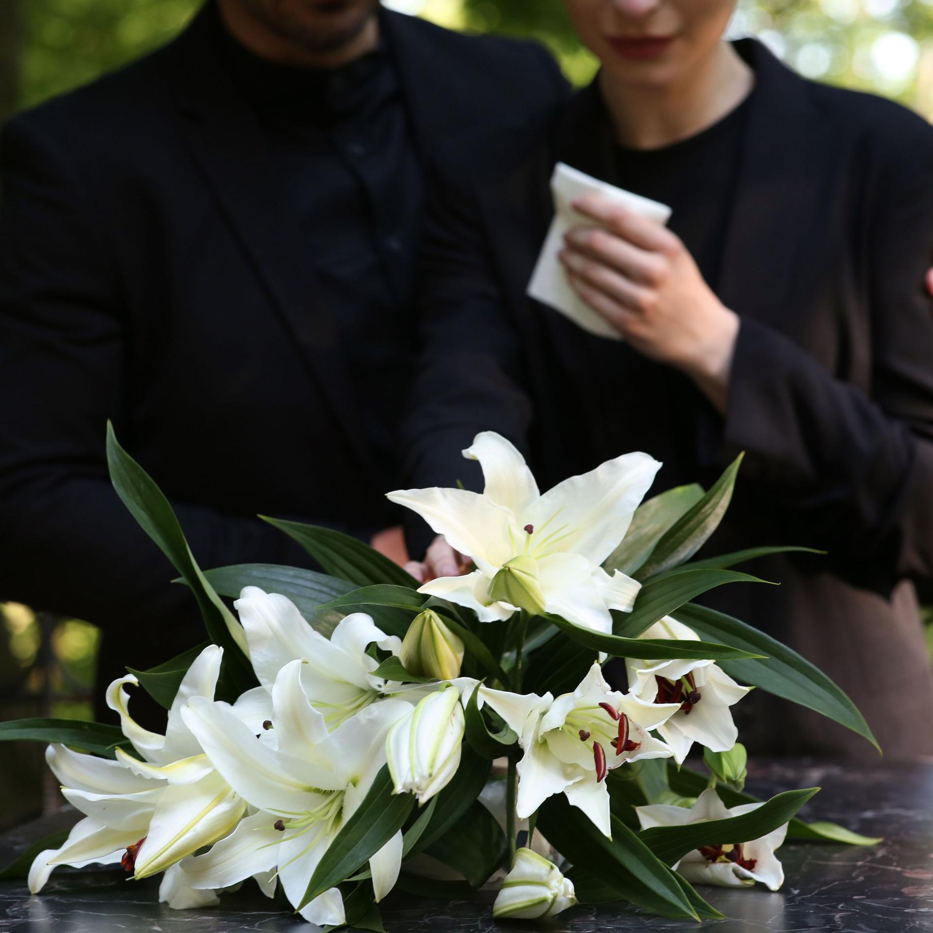 Paar in der Nähe von Granitgrabstein mit weißen Lilien auf dem Friedhof im Freien.