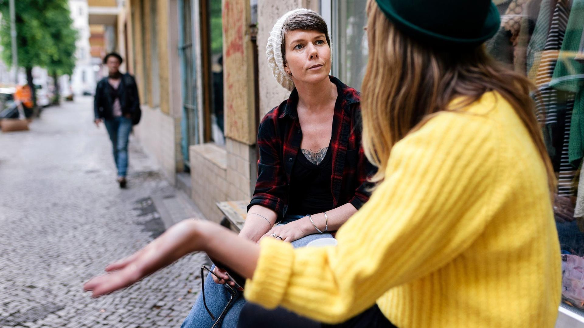 Zwei Frauen sitzen an einem Schaufenster vor einem Laden und führen ein ernstes Gespräch. 
