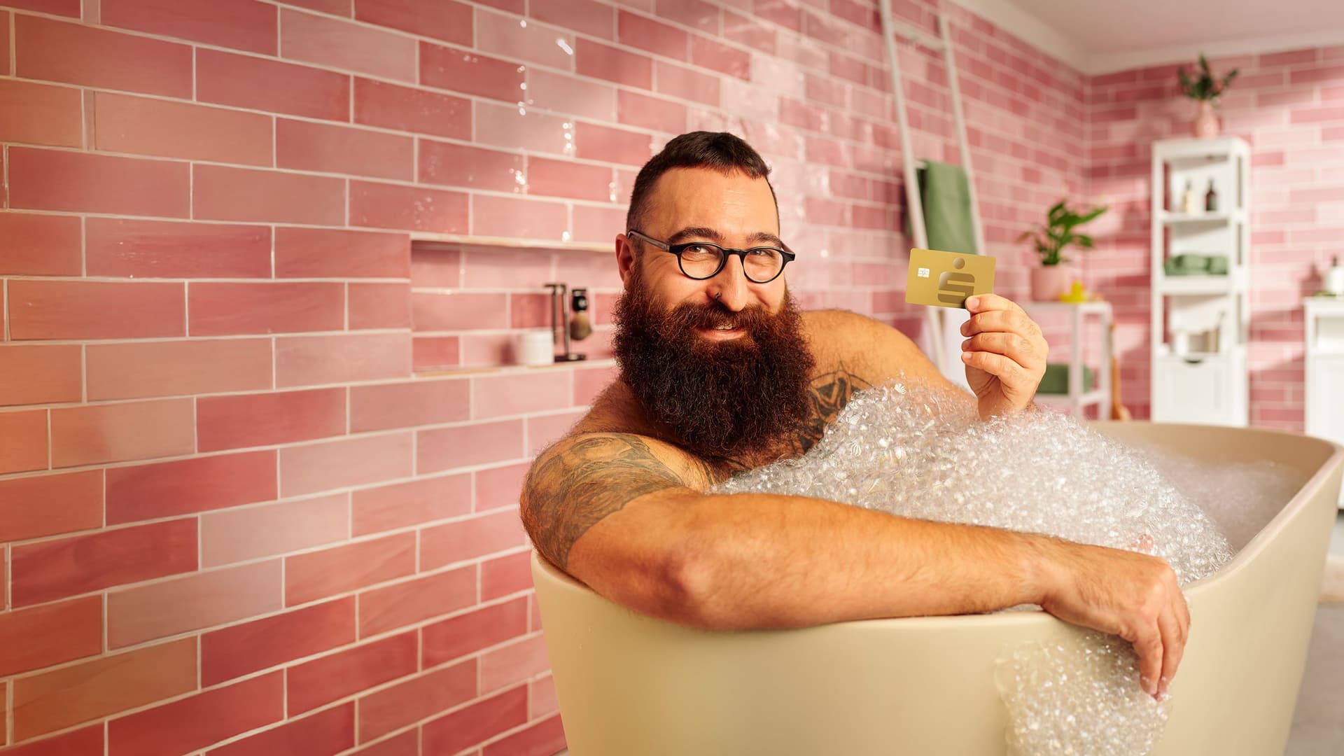 Mann mit Bart und Brille liegt entspannt in einer Badewanne mit Schaum und hält eine goldene Sparkasse-Kreditkarte in die Höhe. Im Hintergrund eine rose-farbene Fliesenwand.