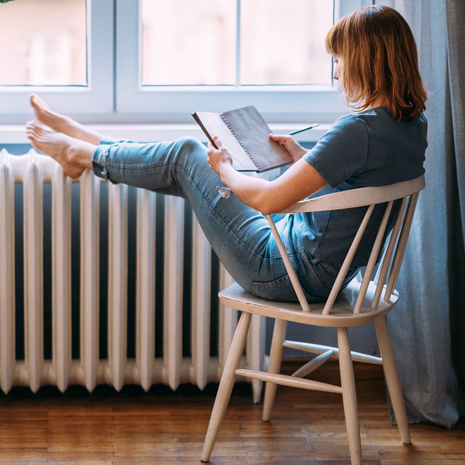 Eine Frau sitzt bequem auf einem Stuhl und liest ein Buch, während sie ihre Füße auf einer weißen Heizung abstützt.
