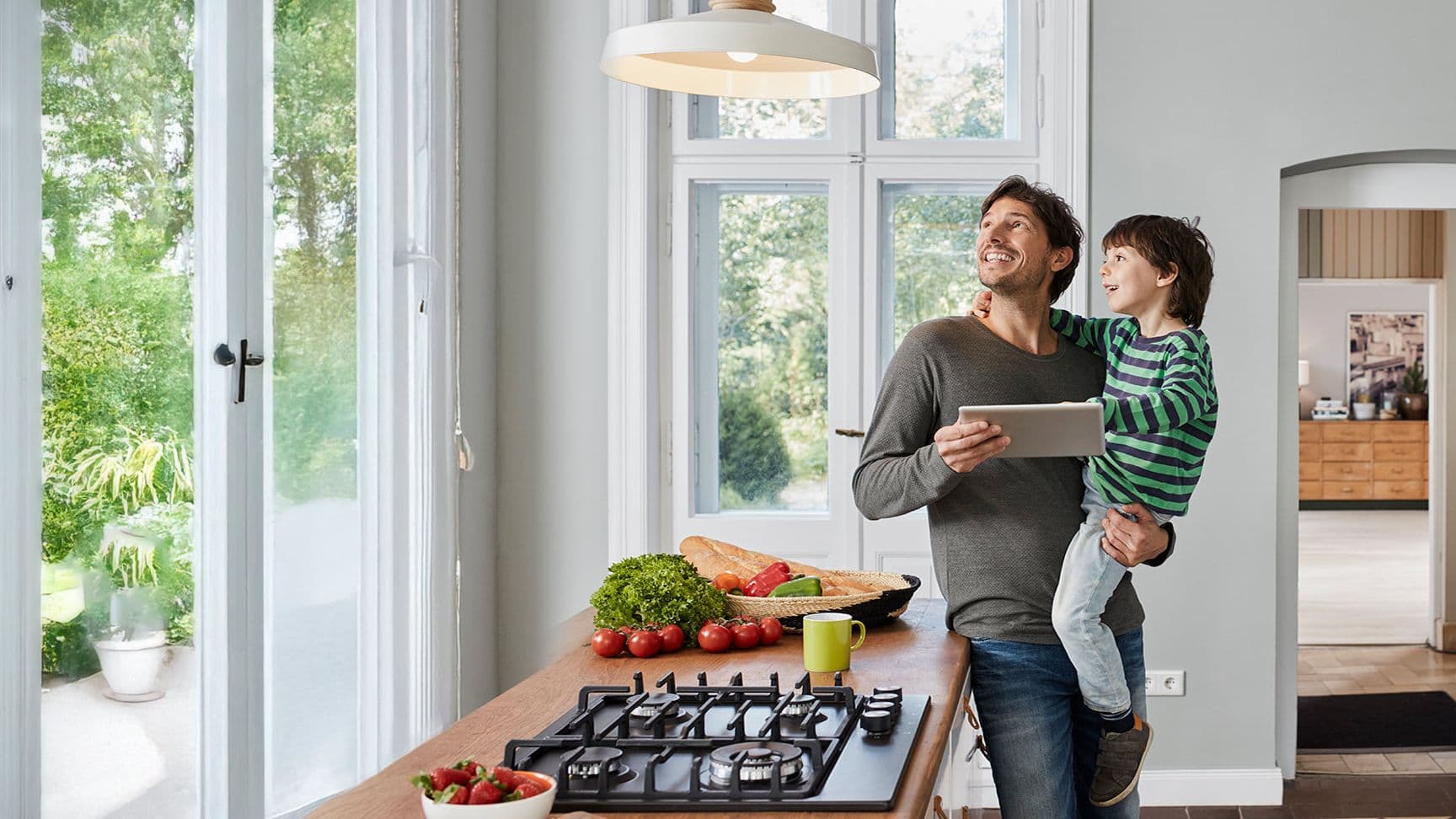 Vater hält Kind im Arm in moderner Küche mit Tablet in der Hand