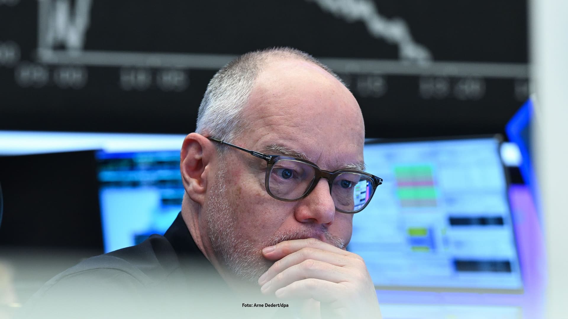 Ein Mann schaut kritisch auf einen Monitor. Im Hintergund ist der fallende Börsengraph zu sehen.