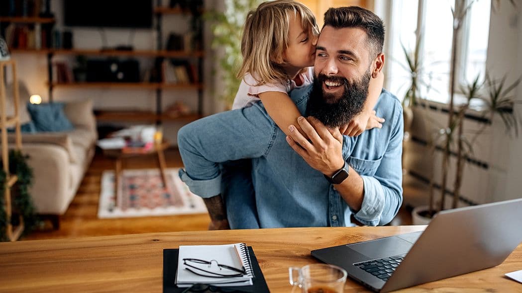 Vater arbeitet am Laptop im Homeoffice, während seine kleine Tochter ihm lachend von hinten um den Hals fällt.