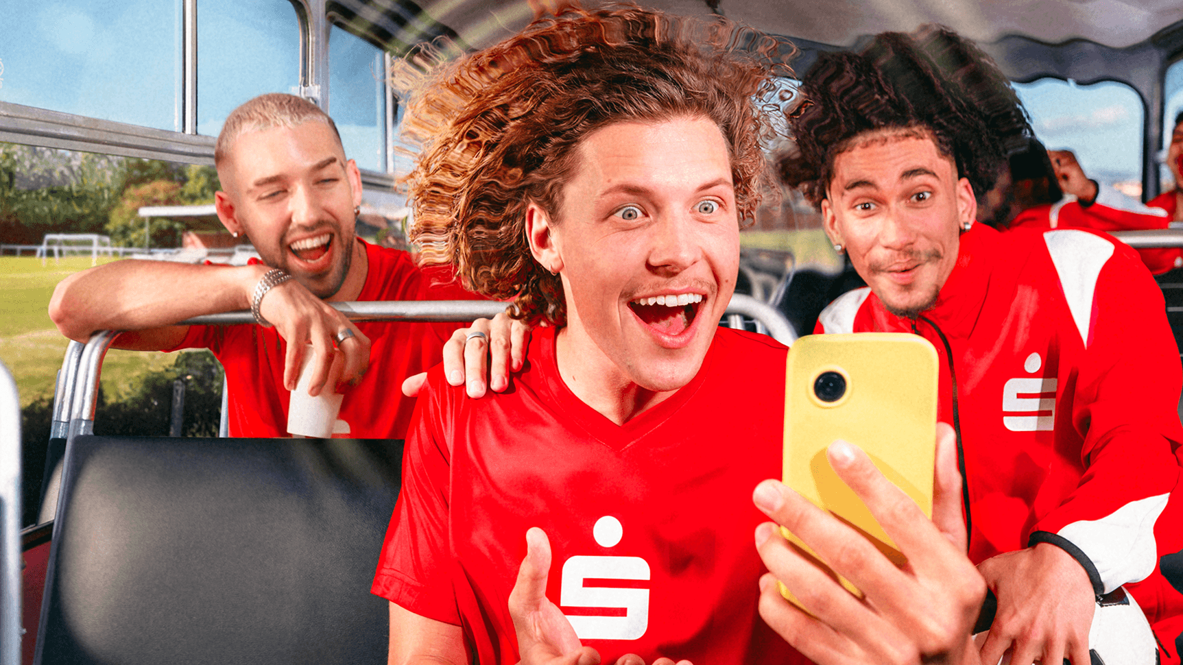 Junge Fußballspieler in roten Trikots sitzen fröhlich in einem Bus; im Vordergrund hält ein Spieler ein gelbes Smartphone