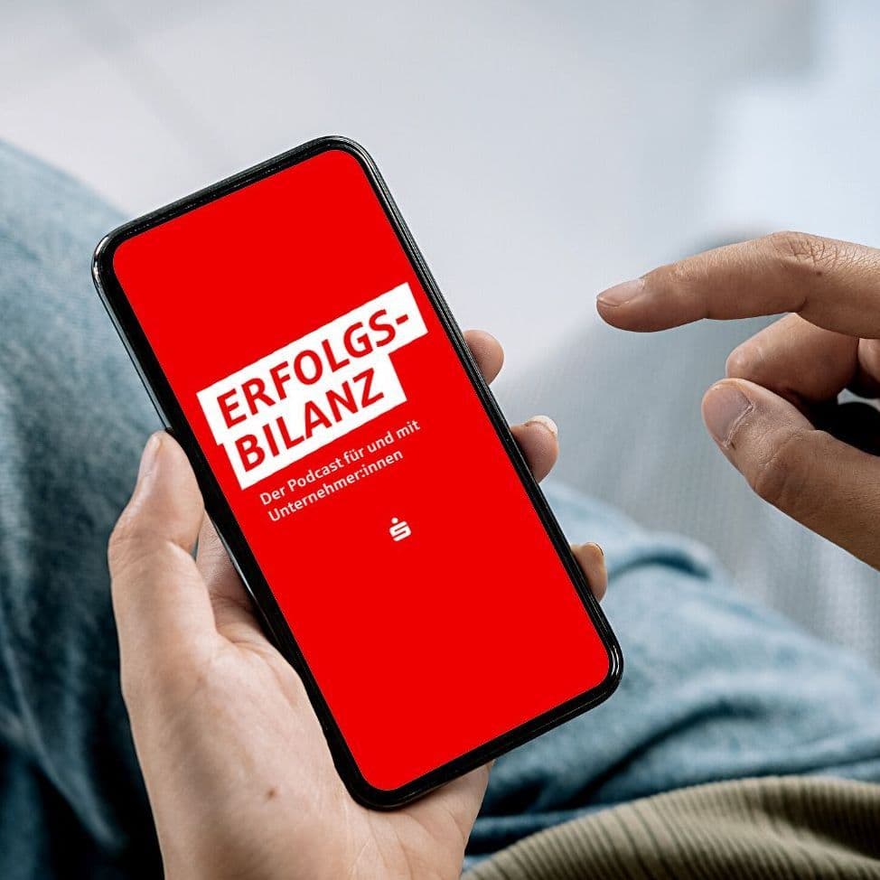 Hand mit Handy, das den Erfolgsbilanz-Podcast anzeigt